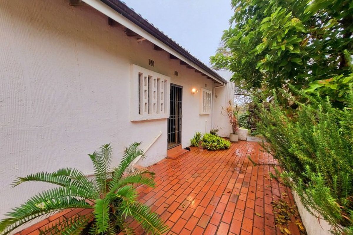 A rear exterior view of the property featuring a brick-paved patio area, security gates, and established garden greenery along the side of the house.
