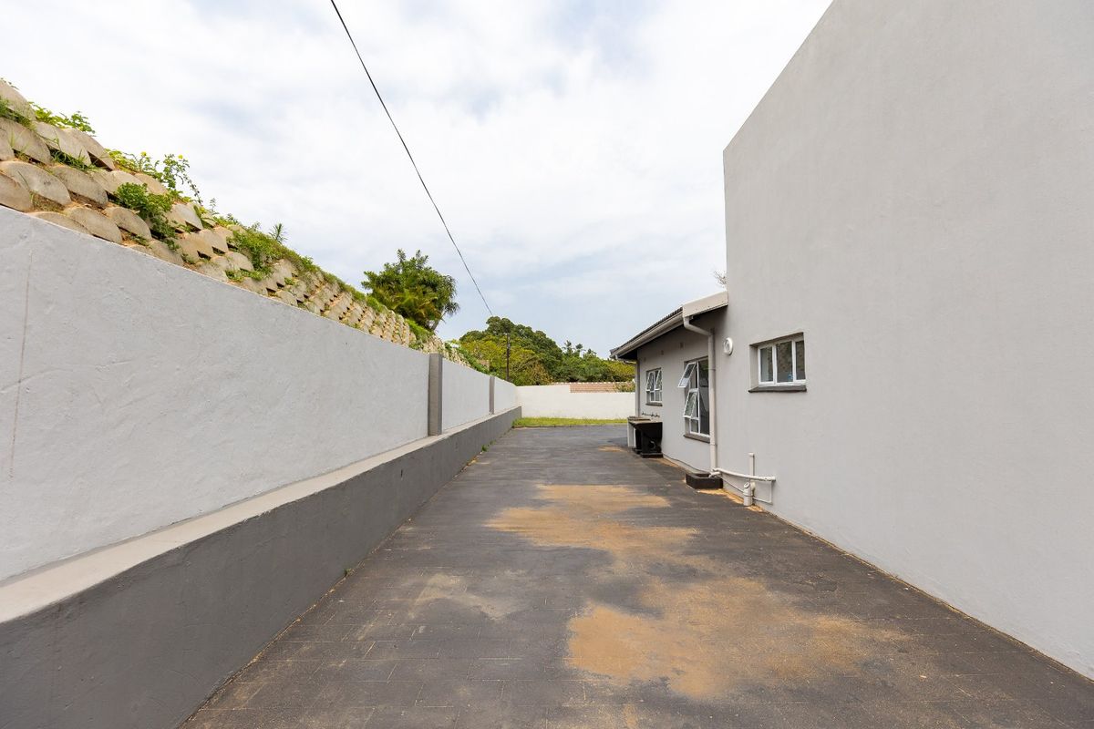 A side exterior view of the property featuring a paved driveway, retaining wall, and exterior building facade with casement windows.
