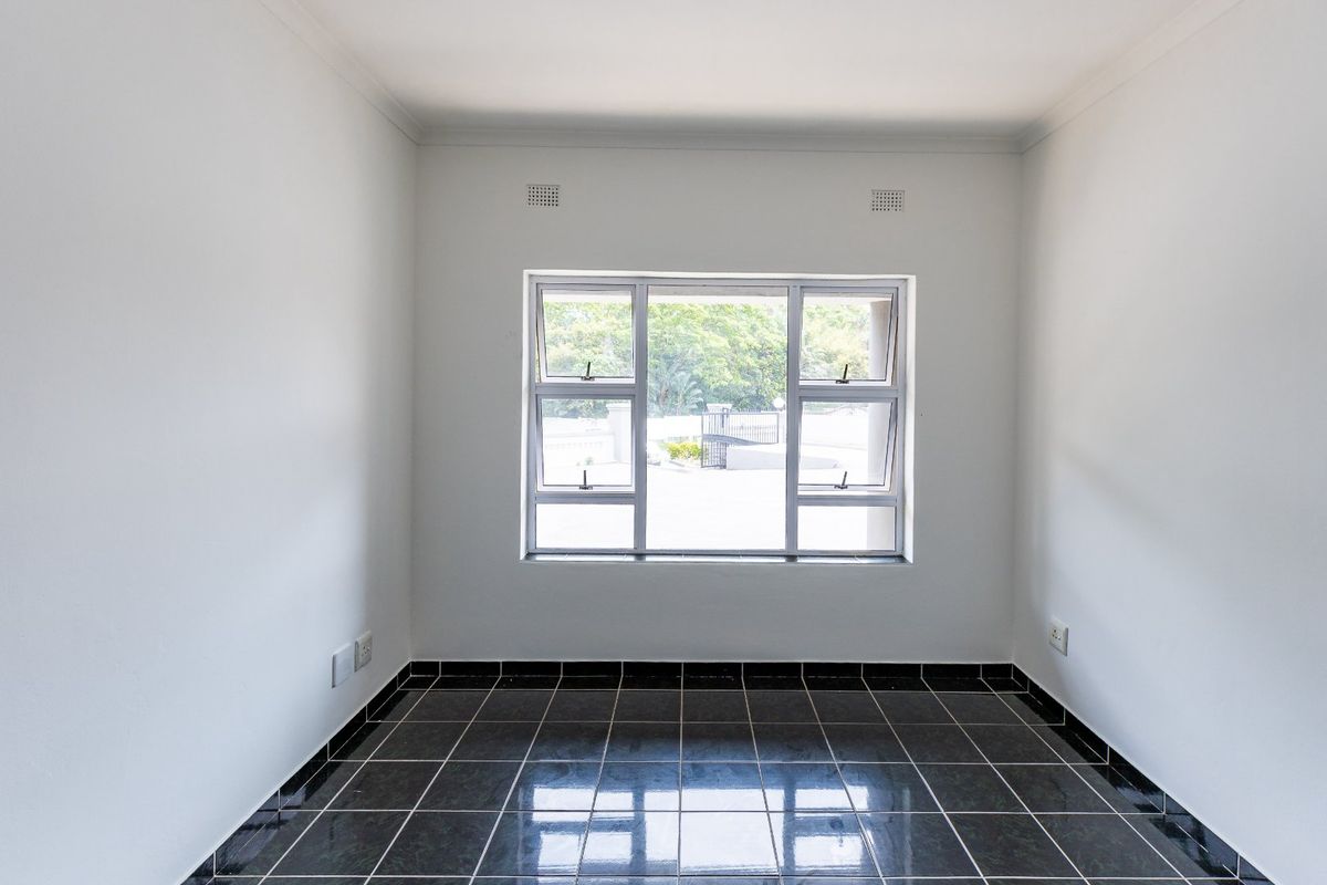A clean, empty bedroom featuring dark tiled flooring, white walls, and a large aluminium window providing natural light.