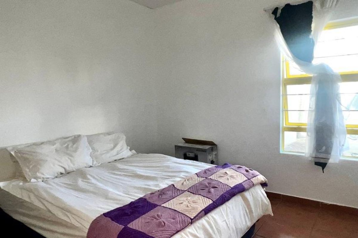 A simple bedroom featuring tiled flooring and a window fitted with security bars, providing natural light to the space.