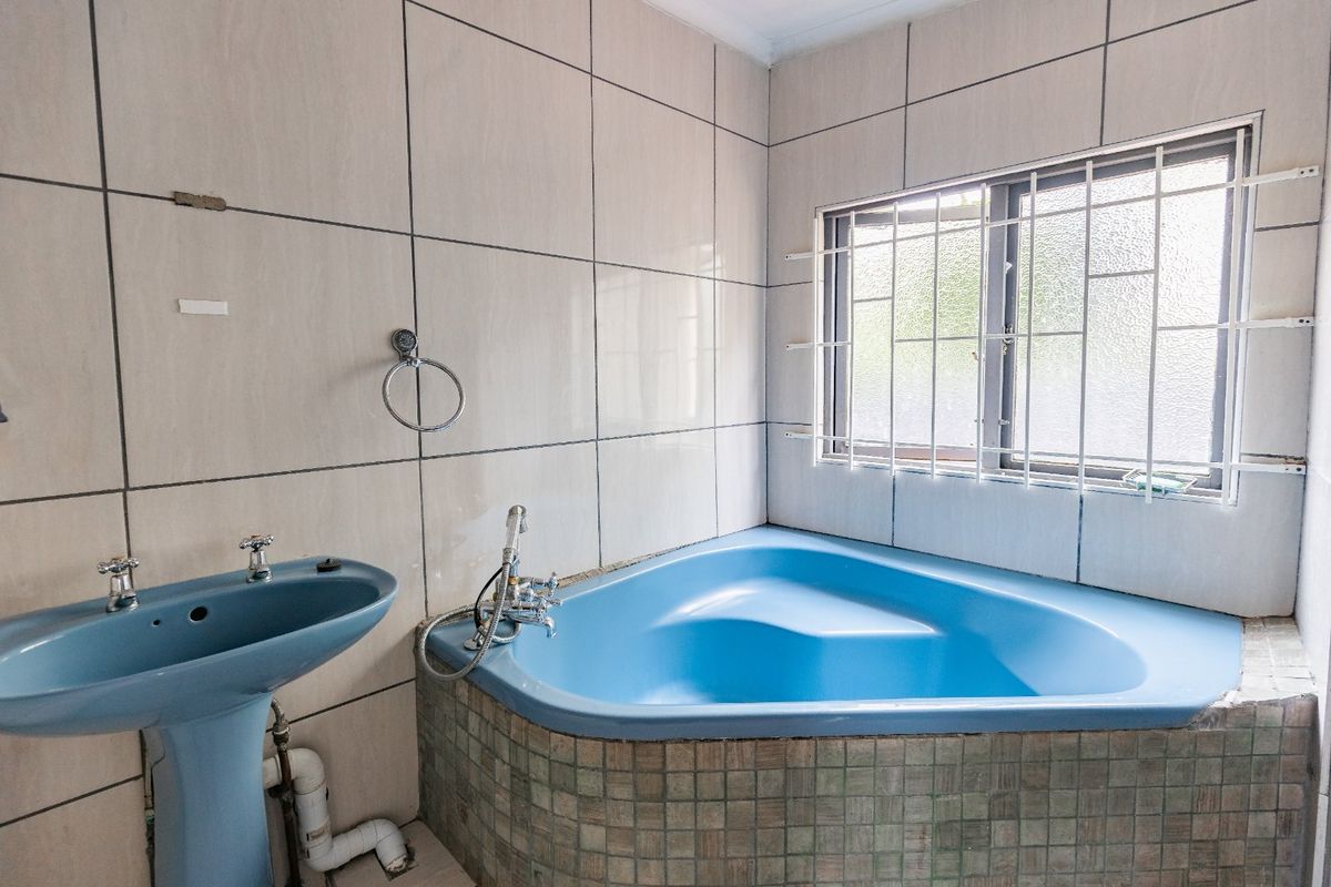A dated bathroom featuring a blue corner bathtub and matching pedestal basin, finished with neutral wall tiles and a window with security bars.