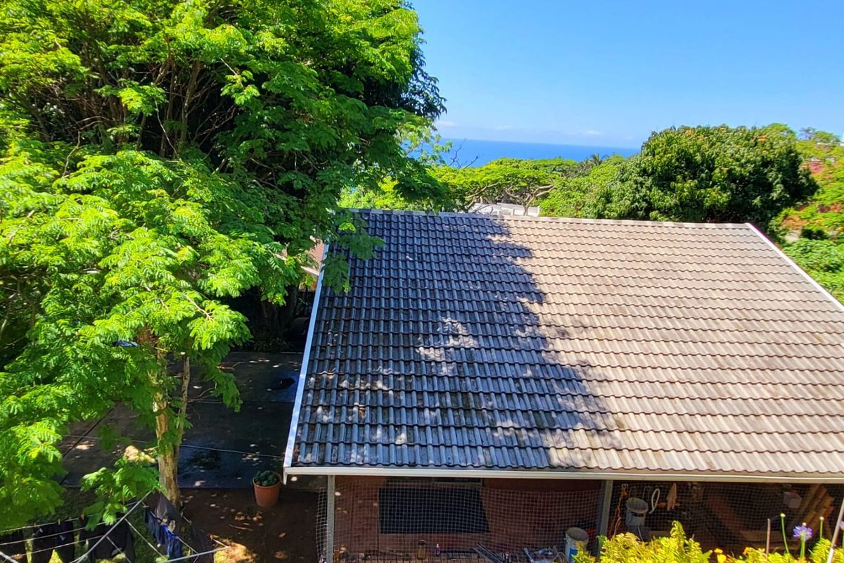 Aerial view of a residential property featuring a tiled roof, brick exterior walls, and surrounding mature trees with a distant ocean view.