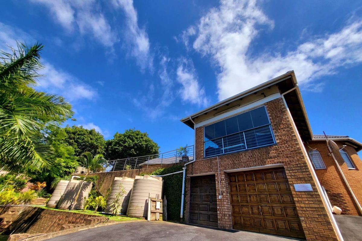 Rear view of the property featuring a brick facade, double garage, elevated balcony with glass balustrade, and multiple water storage tanks.