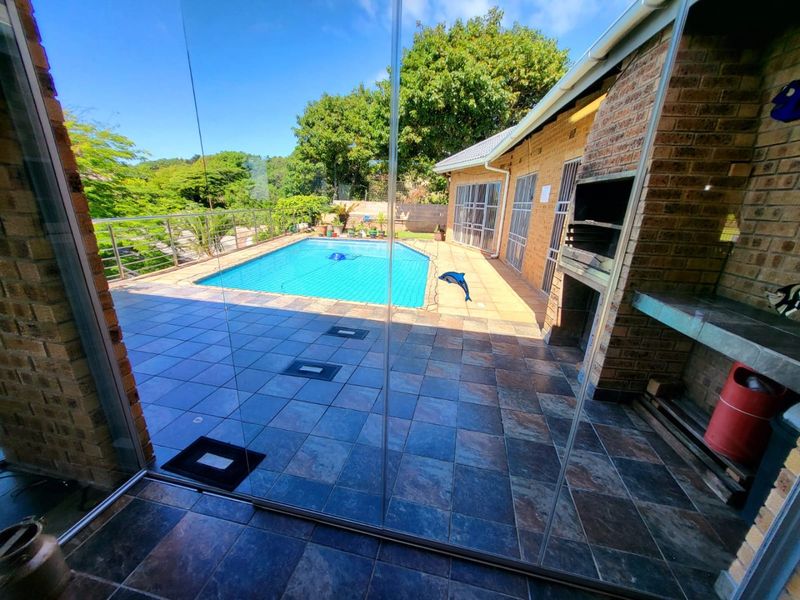 Outdoor entertainment area featuring a tiled patio, built-in braai, and a swimming pool with glass balustrades overlooking the garden.