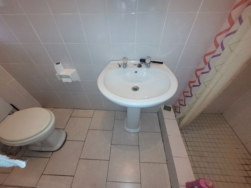 A functional bathroom featuring a pedestal basin, toilet, and shower enclosure with tiled walls and flooring, showing signs of wear and dated finishes.