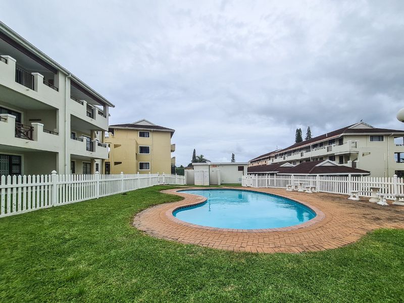 Communal swimming pool area featuring a kidney-shaped pool, brick paving, and white picket fencing surrounded by apartment complex buildings.