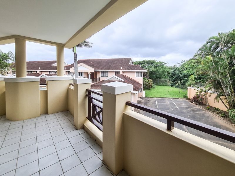 A spacious covered balcony featuring tiled flooring, concrete pillars, and timber railings, overlooking the complex grounds and parking area.