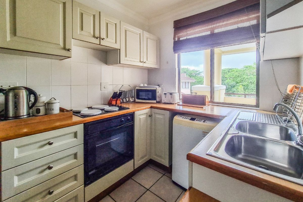 Compact kitchen featuring light-toned cabinetry, wooden countertops, a built-in oven, electric hob, and a double stainless steel sink with a window view.