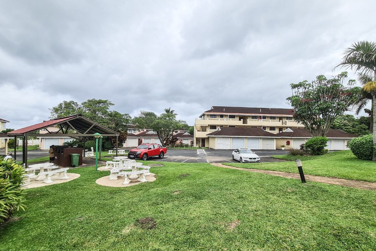 View of the apartment complex exterior featuring a communal garden, covered seating area, and paved driveway with garage access.