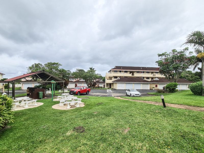 View of the apartment complex exterior featuring a communal garden, covered seating area, and paved driveway with garage access.
