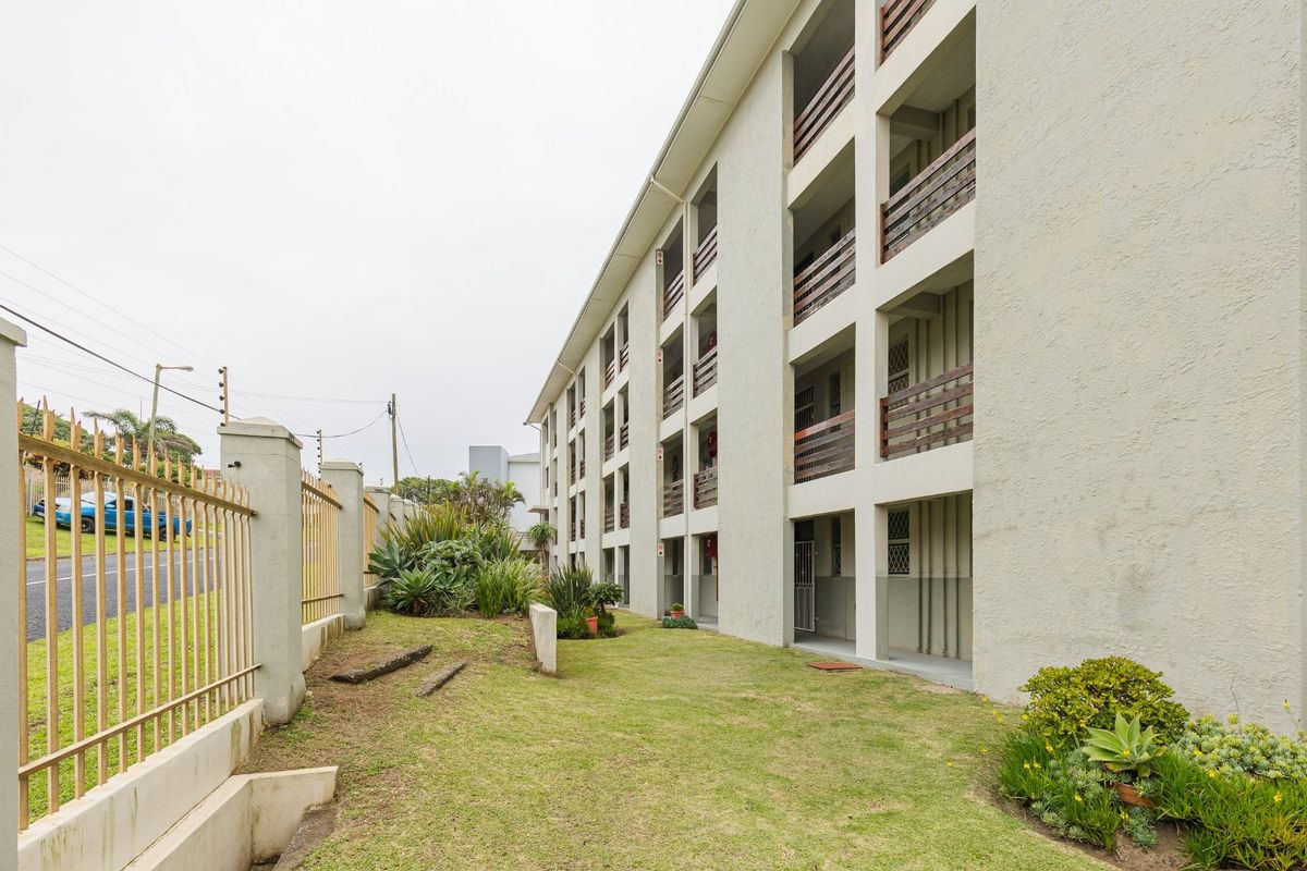 Exterior view of a multi-story apartment building featuring private balconies, concrete structural elements, and a landscaped garden area with perimeter security fencing.