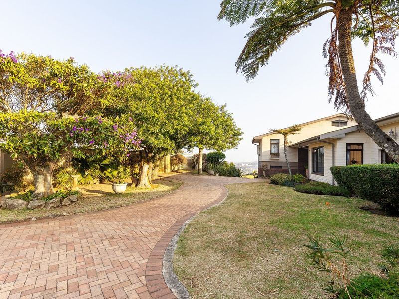 A well-maintained front exterior featuring a curved brick paved driveway, established garden landscaping, and mature trees leading to the main house entrance.