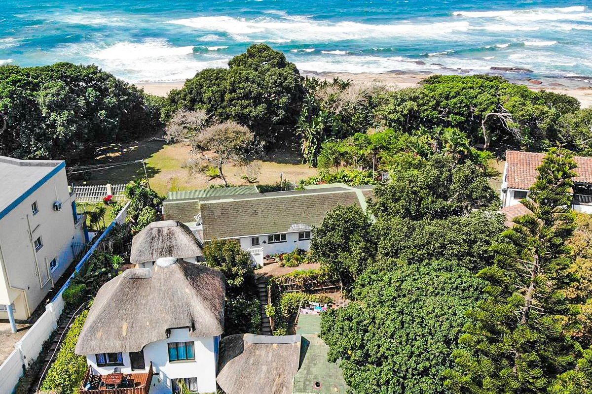 Aerial view of a coastal property featuring multiple structures with thatched and tiled roofs, surrounded by mature trees and proximity to the ocean.
