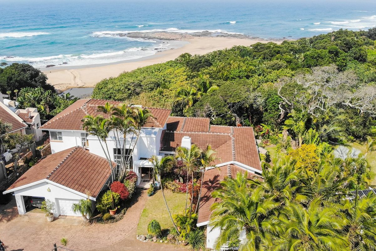 Aerial view of a coastal property featuring a tiled roof, double garage, paved driveway, and lush garden, situated near the beach.