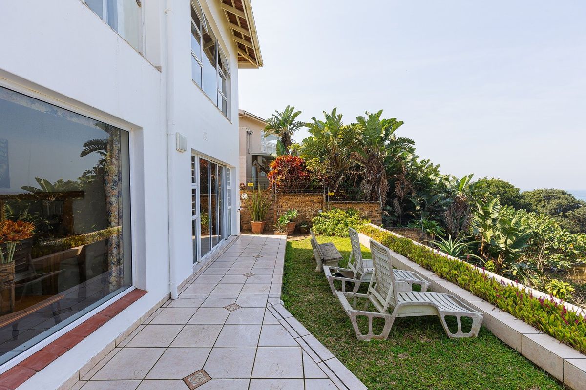 A rear exterior view featuring a tiled patio area, landscaped garden beds, and a retaining wall overlooking the surrounding greenery.