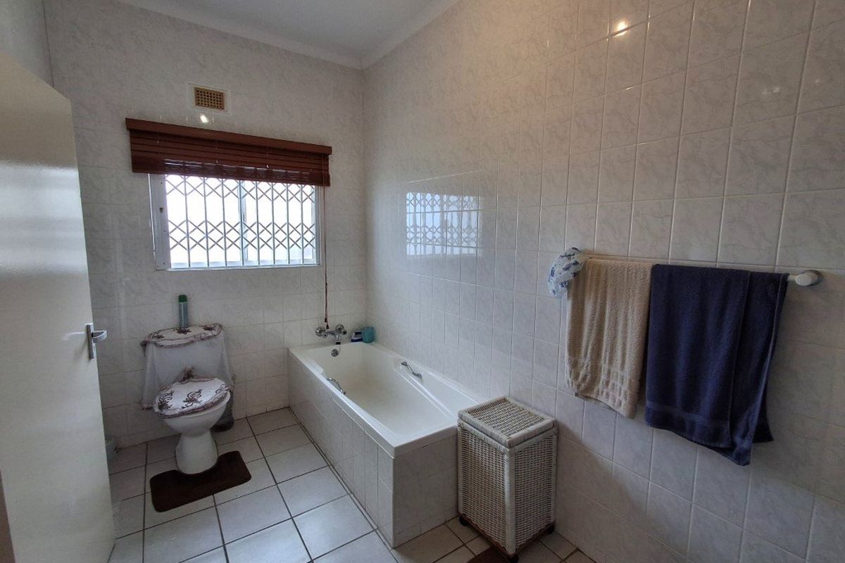 A functional bathroom featuring a built-in bathtub, full-height ceramic wall tiling, tiled flooring, and a window with a security gate.