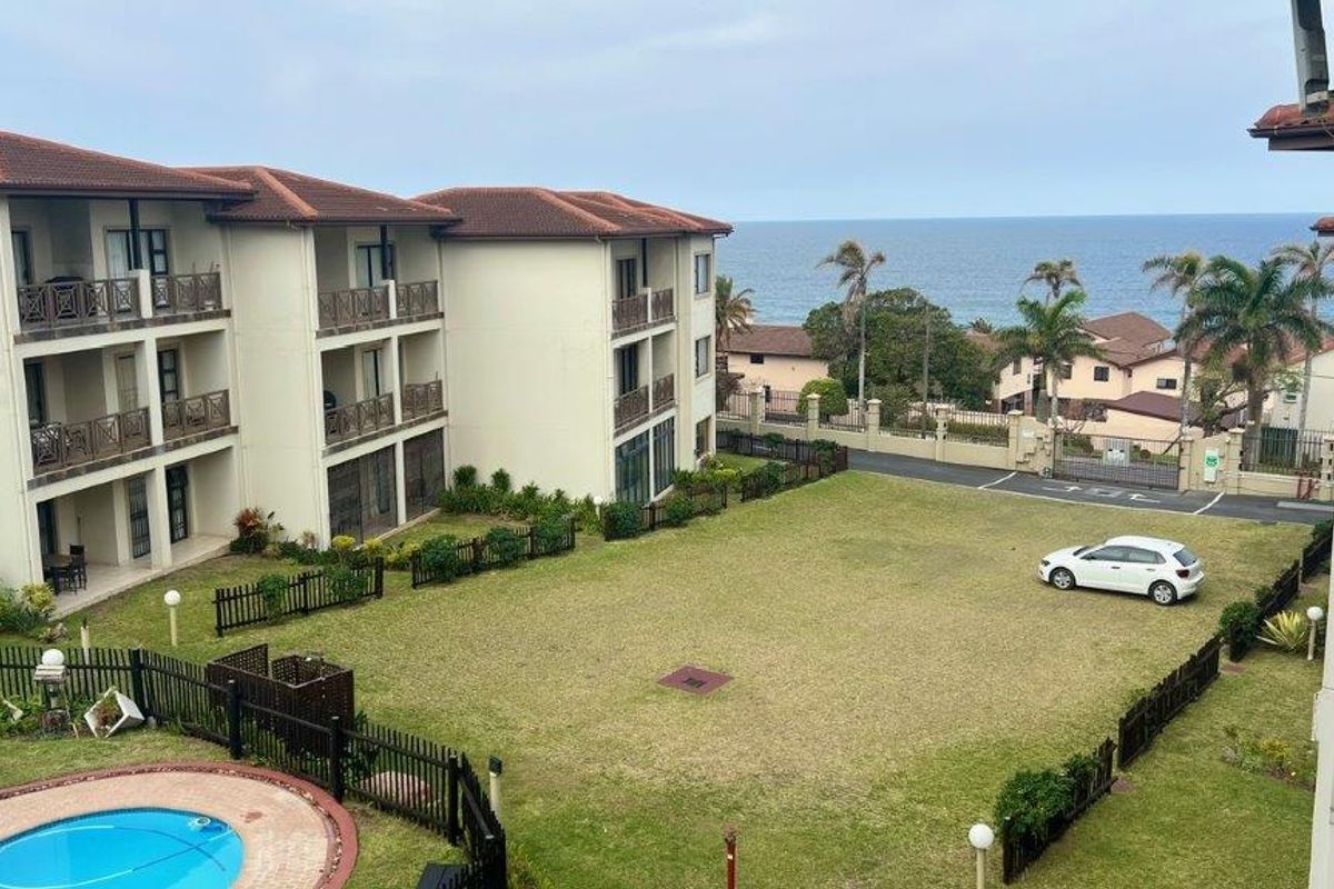 Elevated view of the apartment complex exterior, featuring a communal swimming pool, manicured lawn, and ocean views in the background.