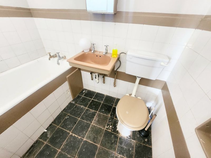 A dated bathroom featuring a wall-mounted basin, standard toilet, and bathtub with original ceramic wall tiling and worn floor finishes.