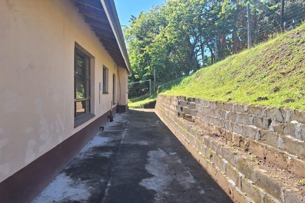 A side exterior view of the property featuring a concrete pathway, a stone retaining wall, and a grass embankment.