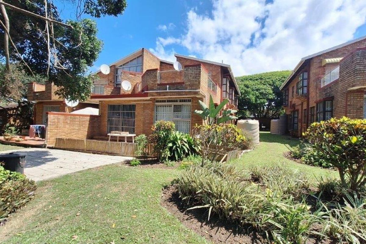 Rear view of a face brick townhouse complex featuring a paved patio area, landscaped garden, and exterior water storage tanks.