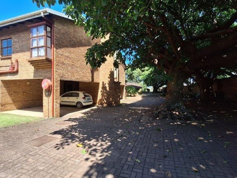 Front exterior view of a face brick townhouse featuring a paved driveway, covered parking area, and mature trees.