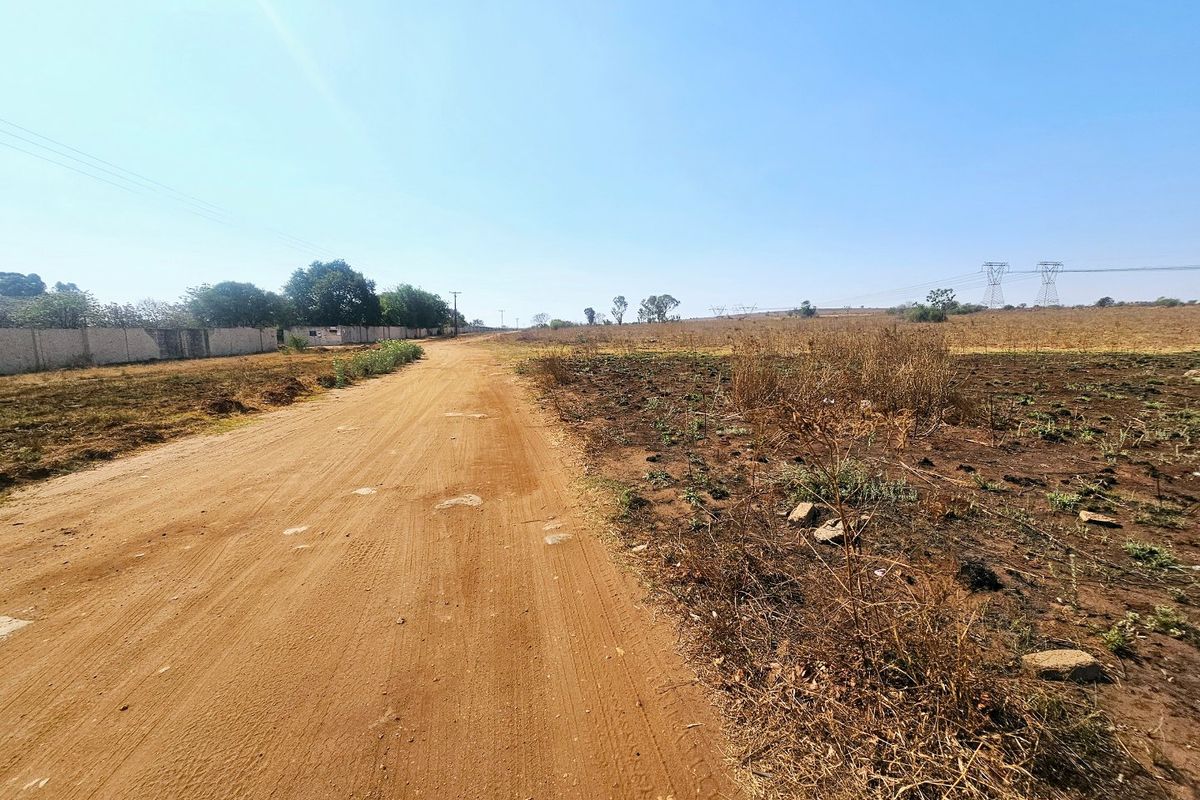 A view of an unpaved dirt road bordering a vacant residential plot with a boundary wall visible in the distance.