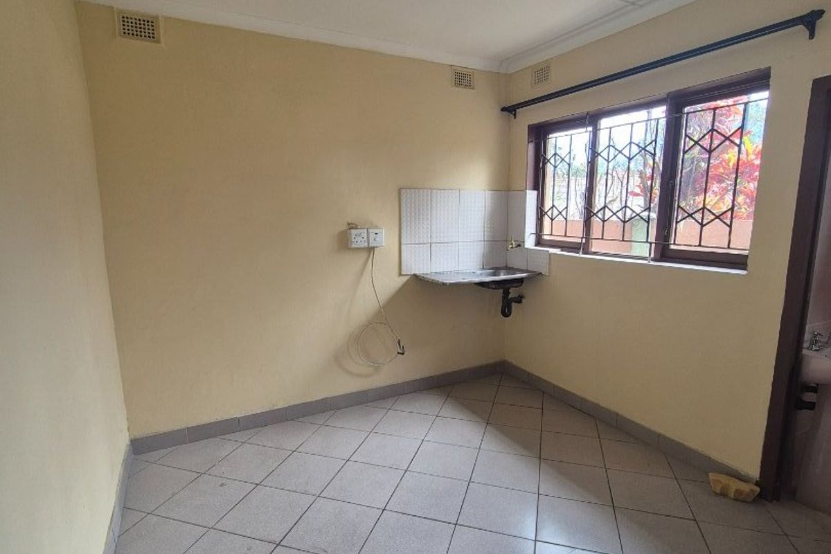 A compact, empty kitchen area featuring tiled flooring, a wall-mounted sink with tiled splashback, and security bars on the window.