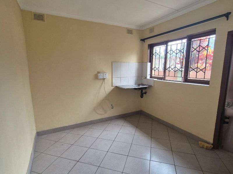 A compact, empty kitchen area featuring tiled flooring, a wall-mounted sink with tiled splashback, and security bars on the window.