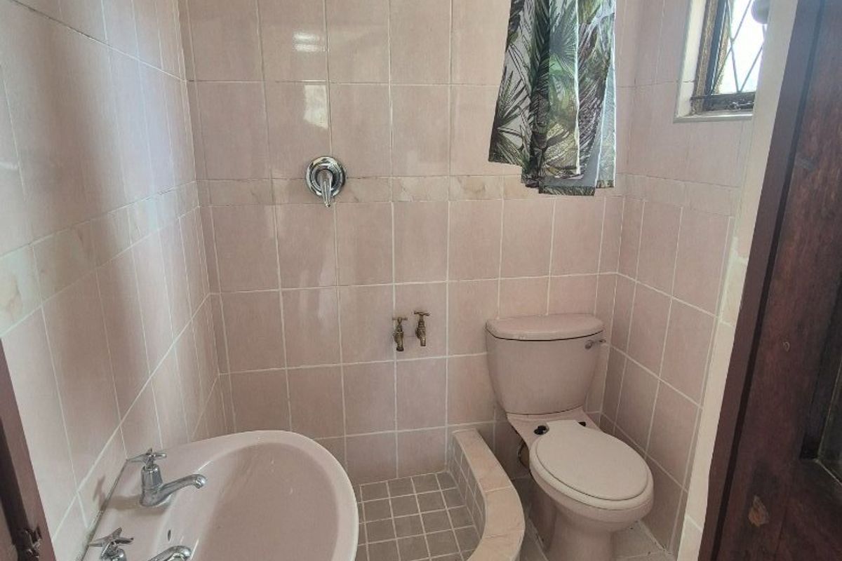 A compact, dated bathroom featuring a pedestal basin, toilet, and a tiled shower area with original pink fixtures and wall tiling.