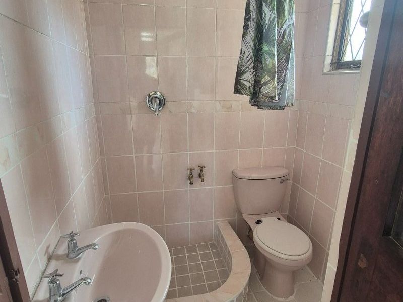 A compact, dated bathroom featuring a pedestal basin, toilet, and a tiled shower area with original pink fixtures and wall tiling.