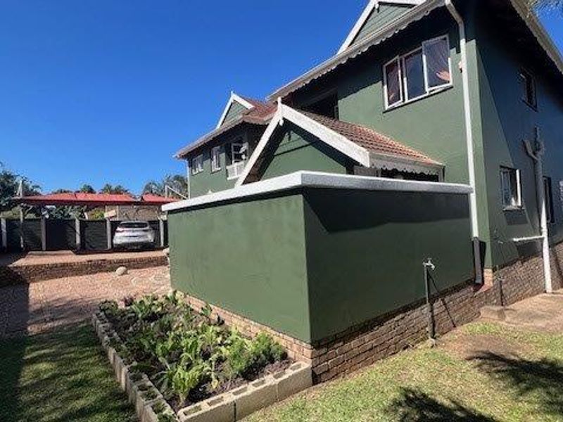 Rear view of a multi-story house featuring a paved courtyard, raised garden beds, and a brick foundation base with ocean views in the distance.