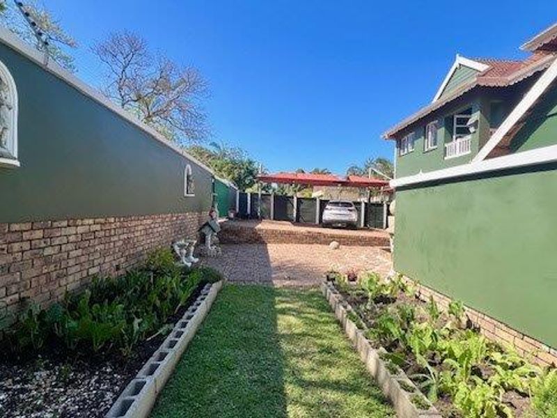 A well-maintained rear exterior area featuring brick boundary walls, raised garden beds, a paved driveway, and a covered carport.