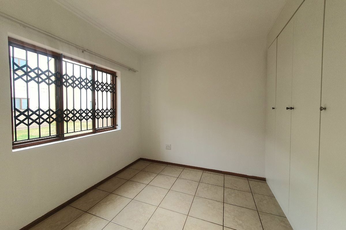 A clean, empty bedroom featuring neutral tiled flooring, built-in cupboards, and a window fitted with a security gate.