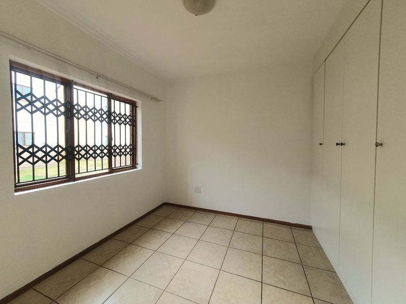 A clean, empty bedroom featuring neutral tiled flooring, built-in cupboards, and a window fitted with a security gate.