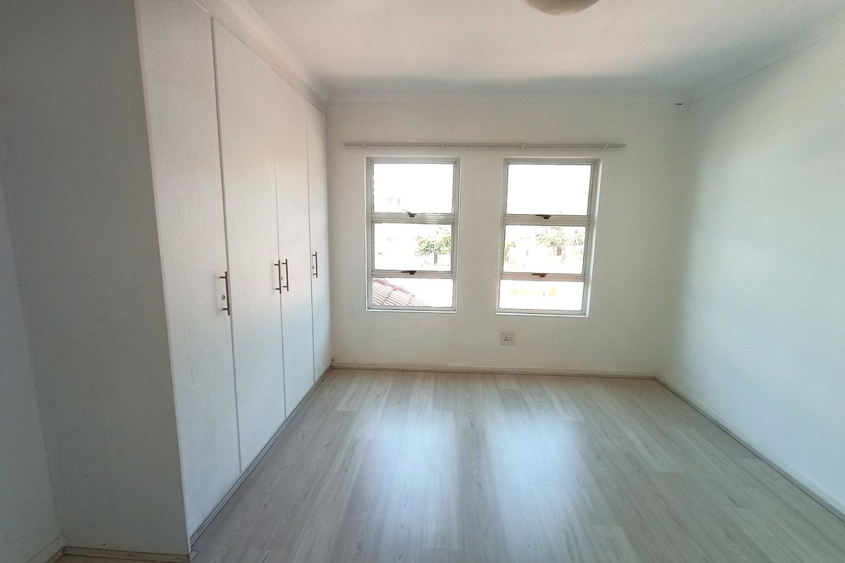 A bright, empty bedroom featuring light-toned laminate flooring, built-in cupboards, and dual windows providing natural light.