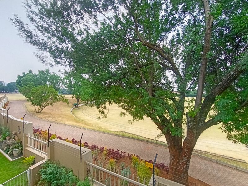 An elevated view overlooking a landscaped park area with mature trees, a paved walkway, and perimeter security fencing.