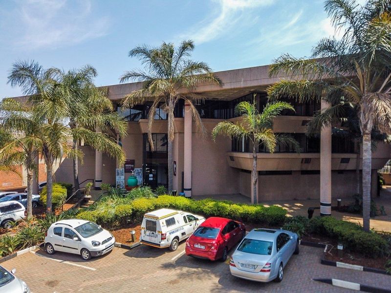 Aerial view of a commercial office building featuring a concrete facade, structural columns, and a paved parking area with landscaped garden beds.