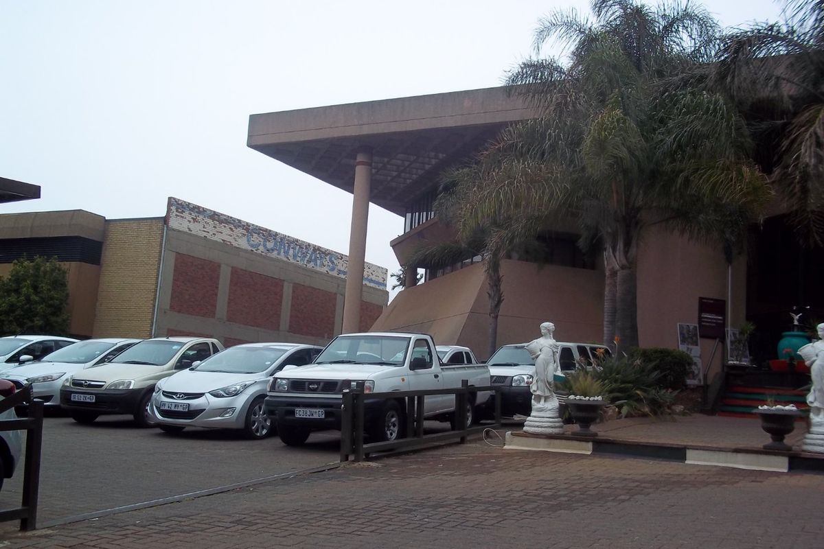 Front exterior view of a commercial building featuring a large overhanging roof structure, concrete pillars, and a paved parking area.