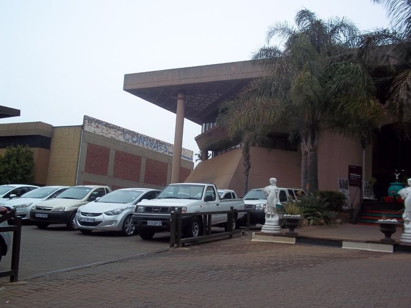 Front exterior view of a commercial building featuring a large overhanging roof structure, concrete pillars, and a paved parking area.