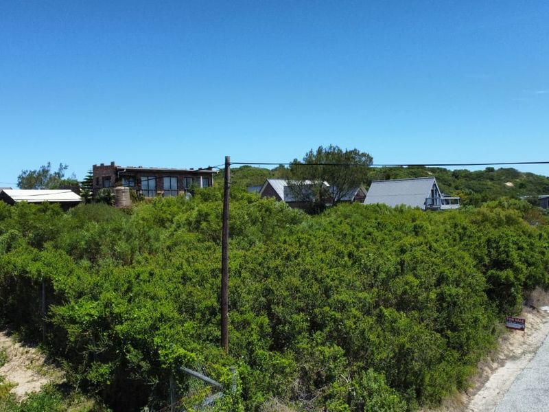 A vacant residential plot covered in dense coastal vegetation, situated along a paved road with surrounding residential properties in a quiet neighborhood.