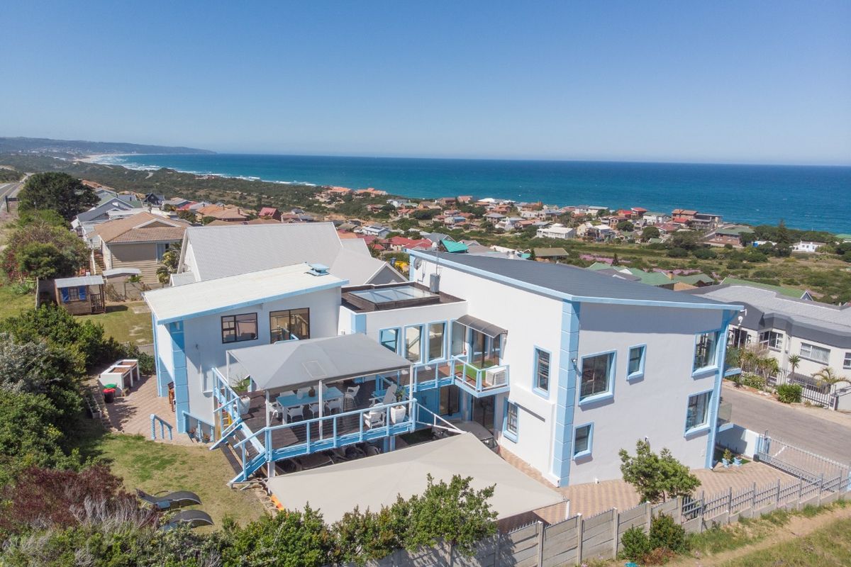 Aerial view of a multi-level coastal residence featuring a large timber deck, covered patio, and paved driveway, set against a scenic ocean backdrop.