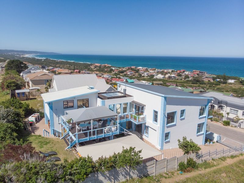 Aerial view of a multi-level coastal residence featuring a large timber deck, covered patio, and paved driveway, set against a scenic ocean backdrop.