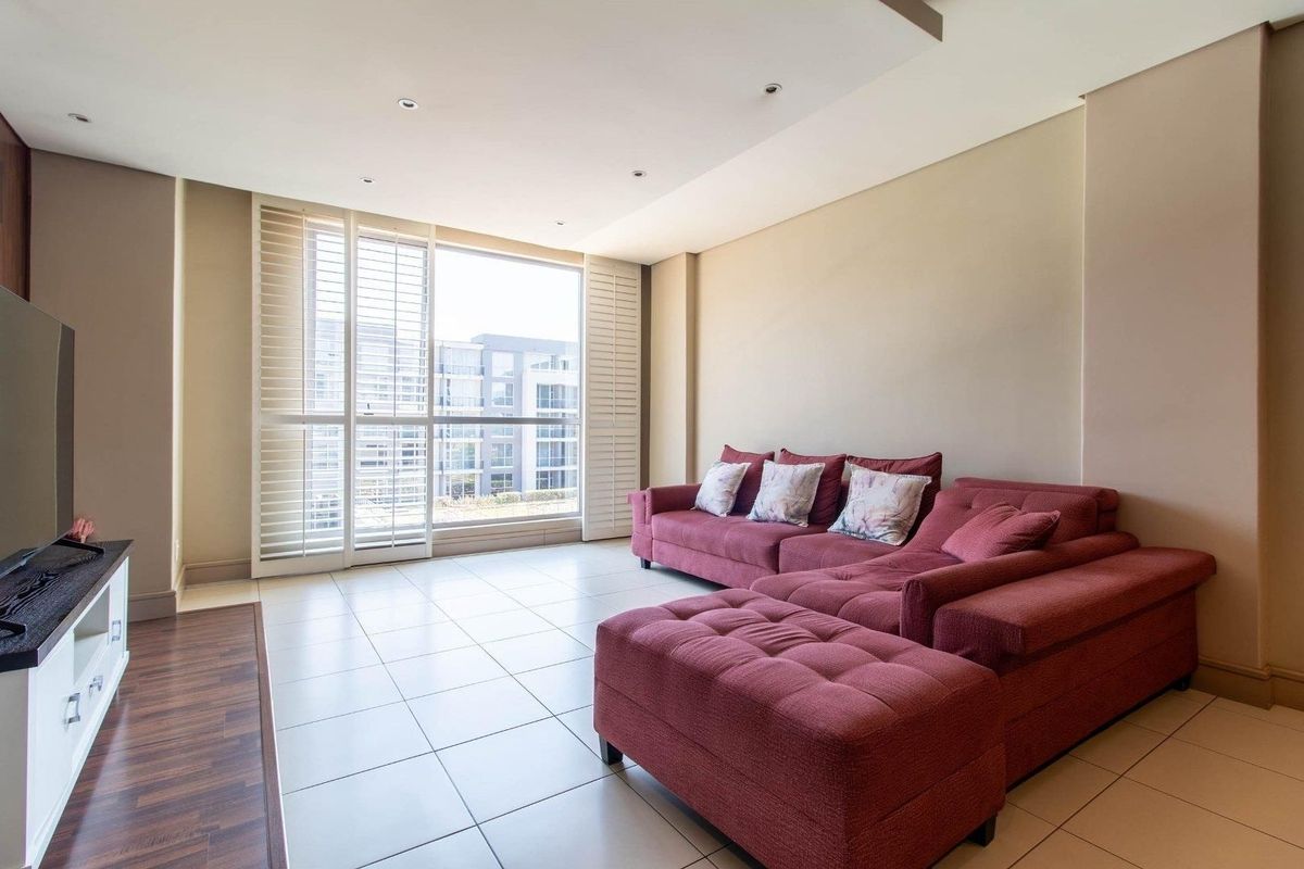 A bright and spacious living room featuring neutral tiled flooring, recessed ceiling lighting, and large windows fitted with plantation shutters.