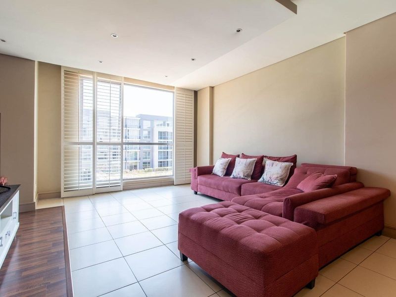 A bright and spacious living room featuring neutral tiled flooring, recessed ceiling lighting, and large windows fitted with plantation shutters.