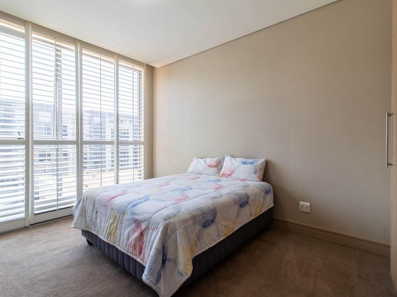 A bright bedroom featuring neutral walls, carpeted flooring, and large windows fitted with plantation shutters, complemented by a built-in wardrobe.