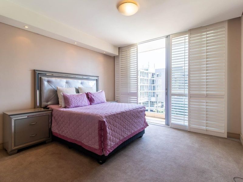 A well-maintained bedroom featuring neutral carpeting, recessed lighting, and plantation shutters opening onto a private balcony.