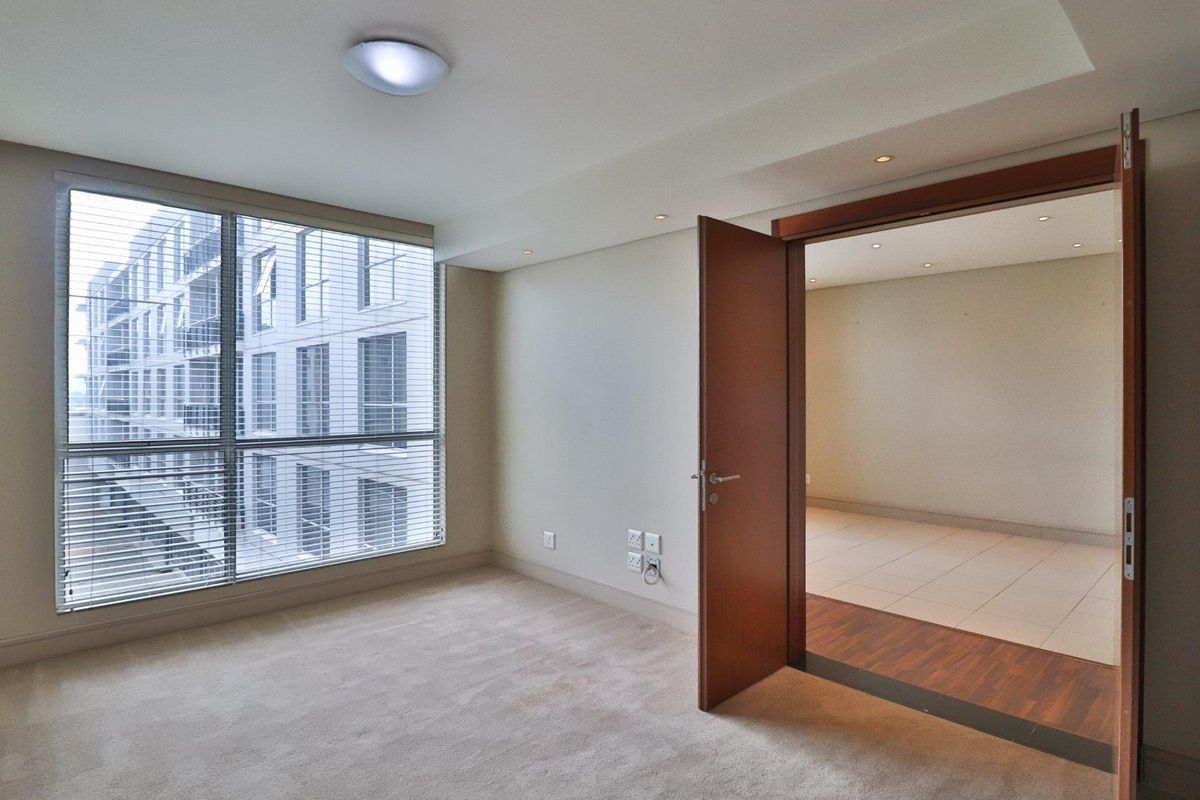 A bright, empty bedroom featuring neutral carpeting, large windows with blinds, and recessed lighting, with an adjacent tiled room visible through an open doorway.