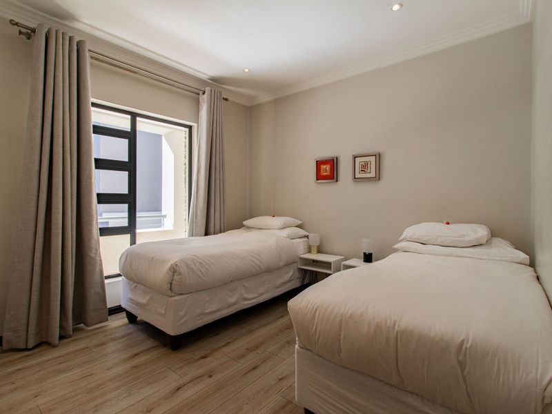 A well-maintained bedroom featuring light-toned laminate flooring, recessed ceiling lighting, and a large window with neutral curtains.