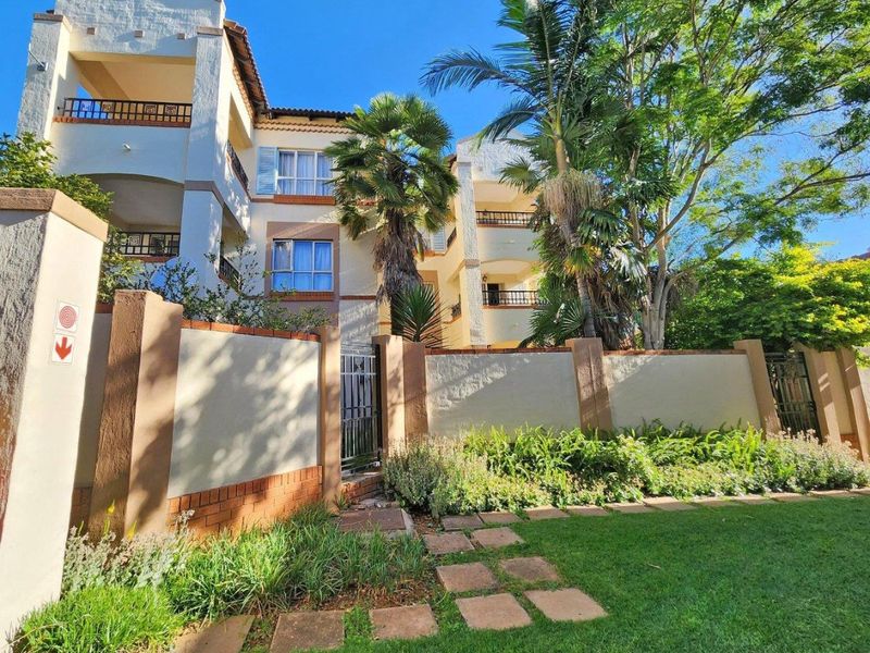 A well-maintained apartment complex exterior featuring a perimeter wall, landscaped garden, paved walkway, and multiple balconies with wrought iron railings.
