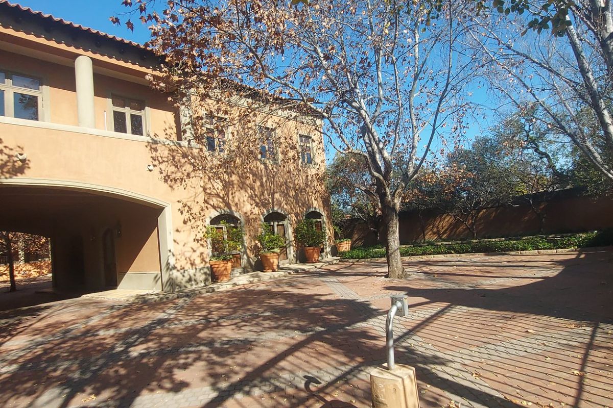 A spacious paved courtyard area featuring a covered walkway, terracotta-toned building facade, and mature trees, providing a well-maintained outdoor space.